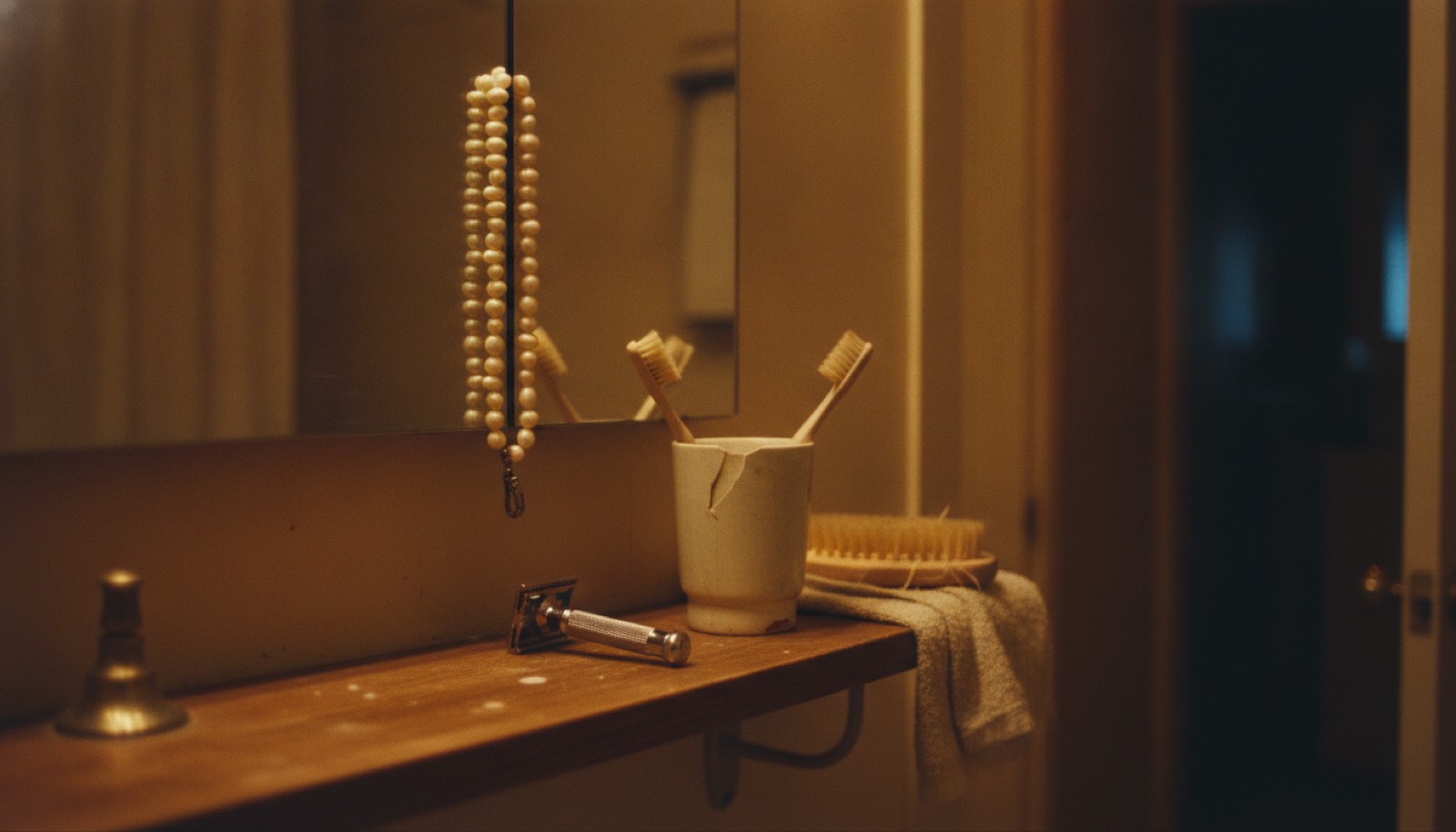 A strand of cream pearls hung on the corner of a bathroom mirror above a wooden shelf holding two well-used toothbrushes in a chipped cup, a heavy razor and a worn hand towel under one warm bulb at night