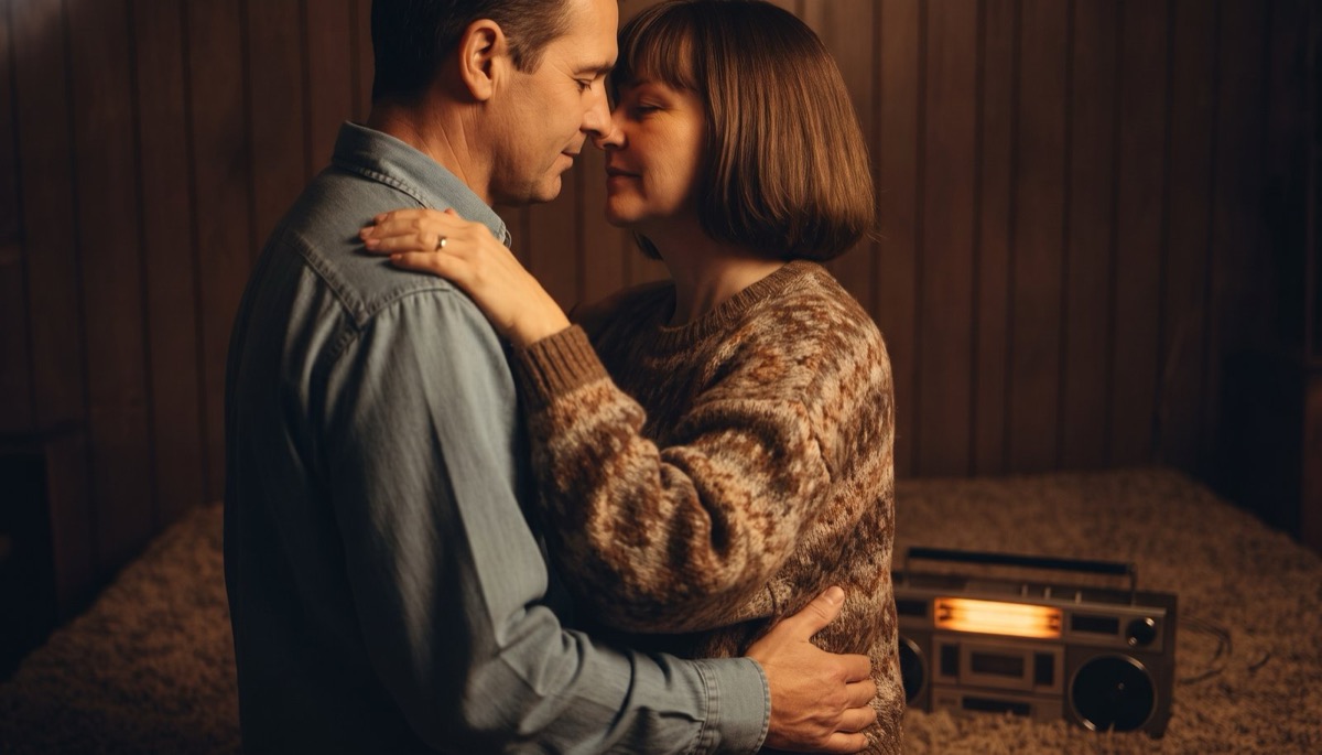 A husband's hand holding his wife's as the two of them stand close together on a wood floor in warm lamplight