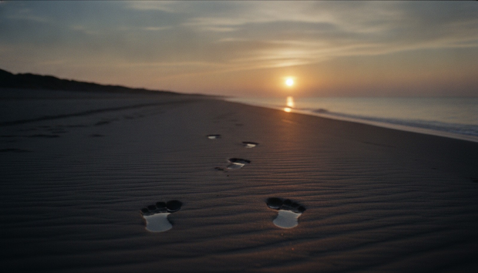 An empty stretch of wet sand at sunrise with two sets of footprints leading toward the water