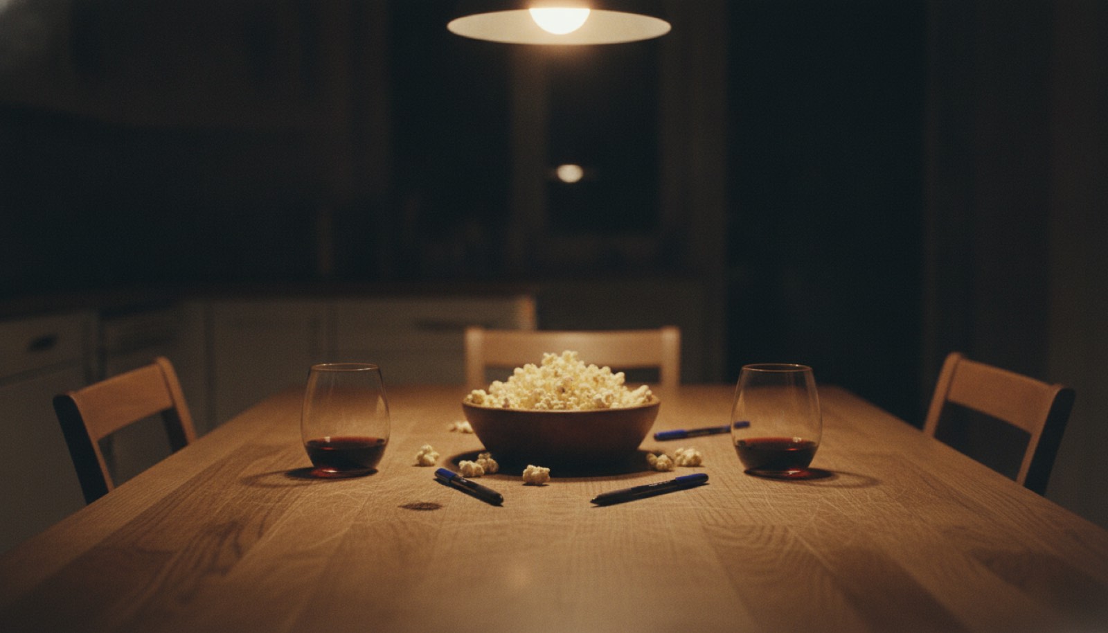 Two stemless wine glasses, a bowl of popcorn and two ballpoint pens on a scratched kitchen table under a warm pendant light
