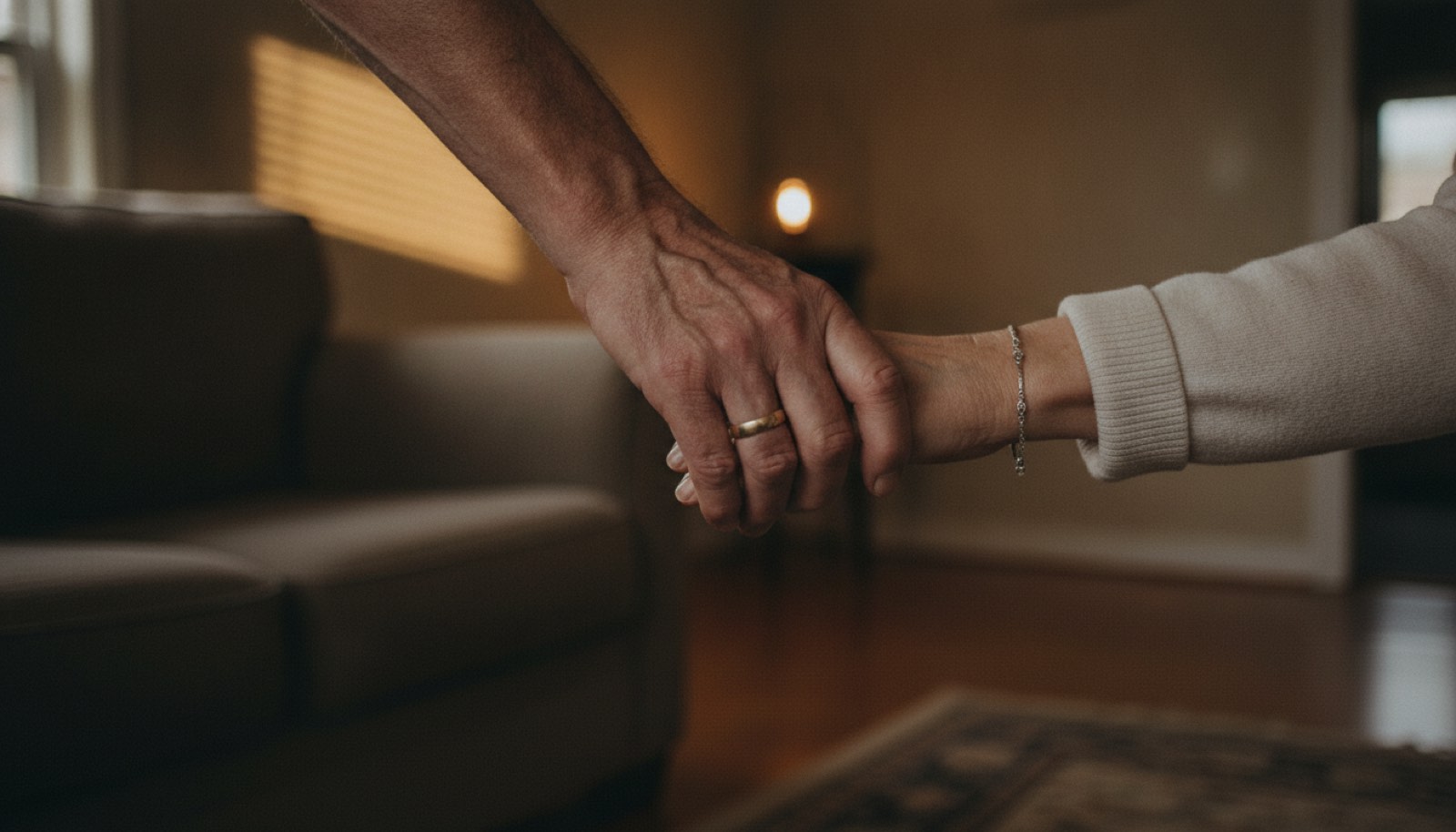 A close view of an older couple's joined hands, his clasping hers, worn wedding bands on both, in a living room lit by low late afternoon window light with the arm of a couch and the edge of a rug behind them