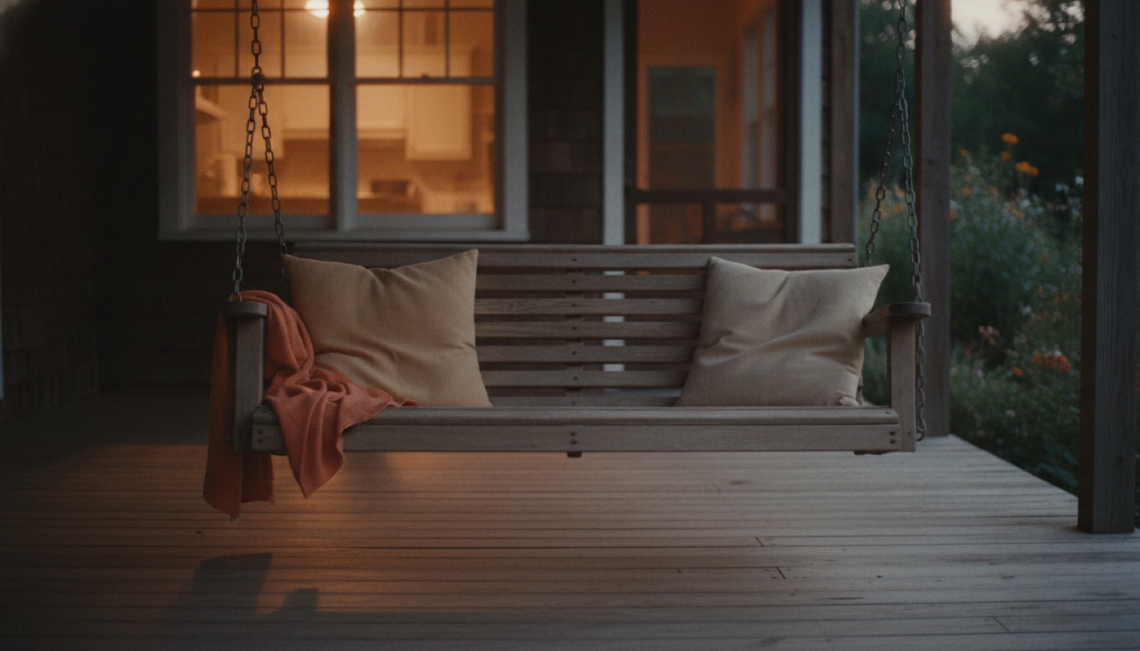 A two seat porch swing hanging still on its chains at dusk, one cushion dented and one untouched, with warm light from the kitchen window falling across the porch boards