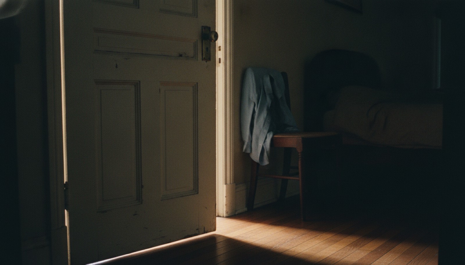 A half open bedroom door with warm light in the hallway beyond and a folded shirt on a chair
