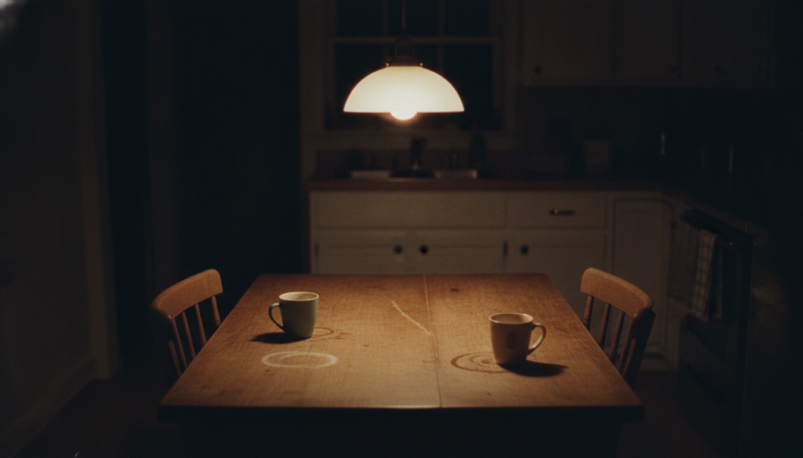 A kitchen table with two chairs pulled in and a single lamp lit above it late at night