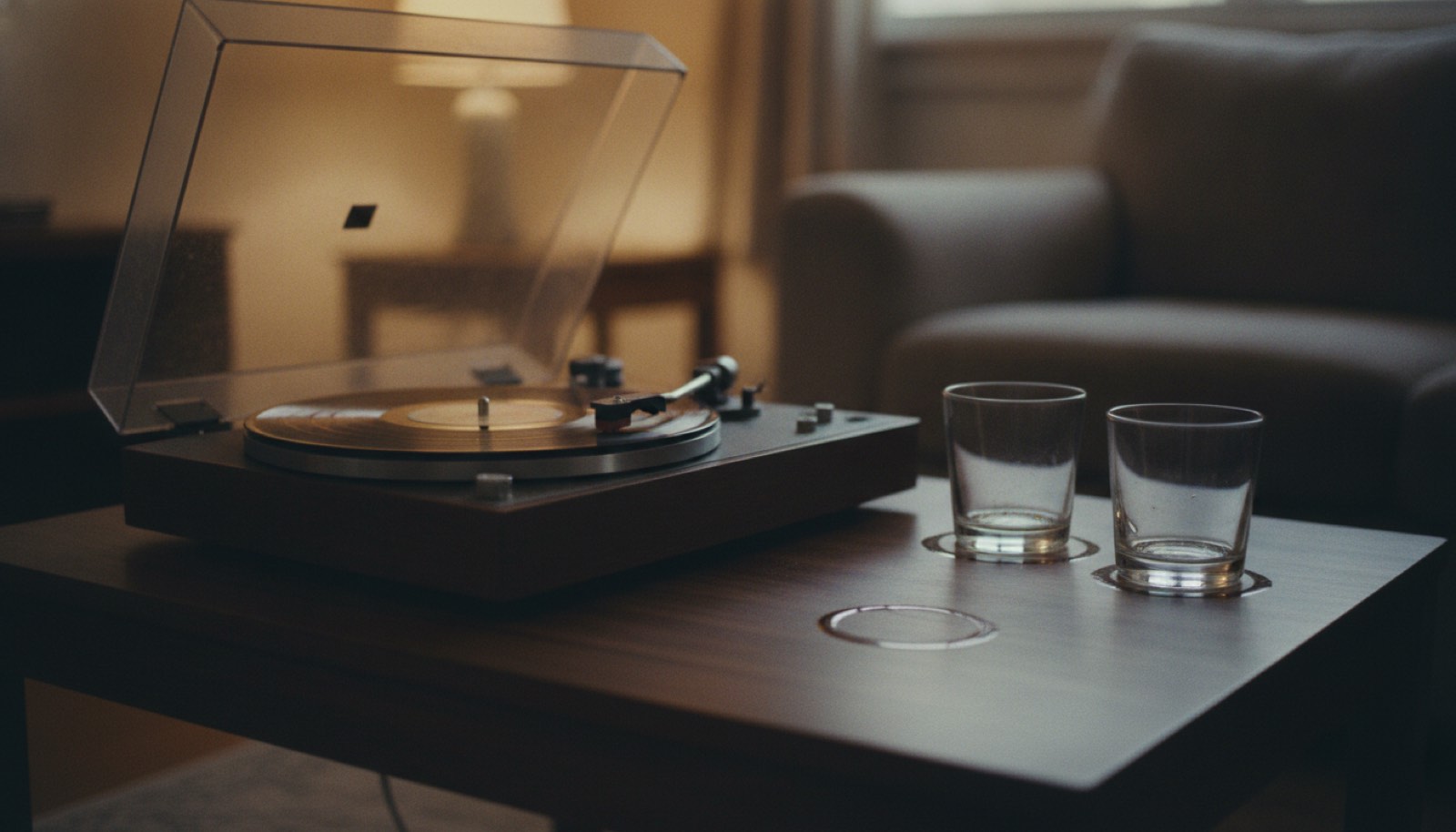 A record player and two glasses on a low table in a living room lit by one lamp