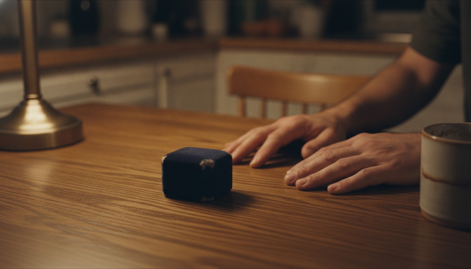 A closed ring box sitting on a kitchen table beside a man's hands under warm evening lamplight