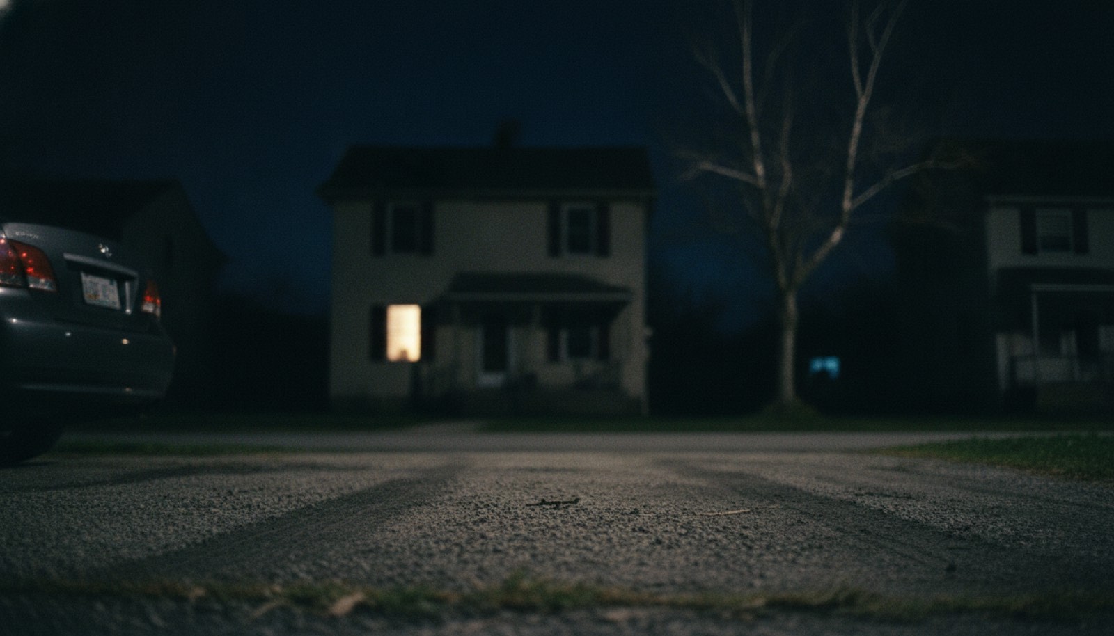 A single lit window in a dark house seen from the end of a driveway at night