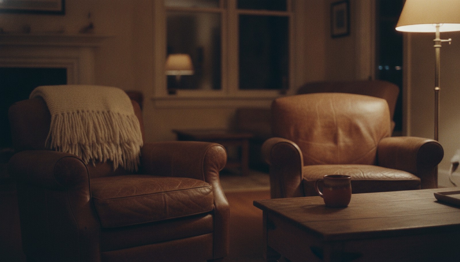 Two worn armchairs facing each other across a low table in a lamplit living room in the evening