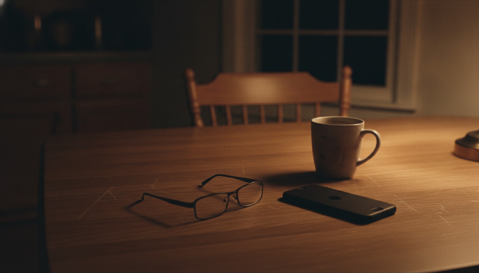 A kitchen table late in the evening with a phone lying face down beside a half-finished mug of coffee and a pair of reading glasses under a warm lamp