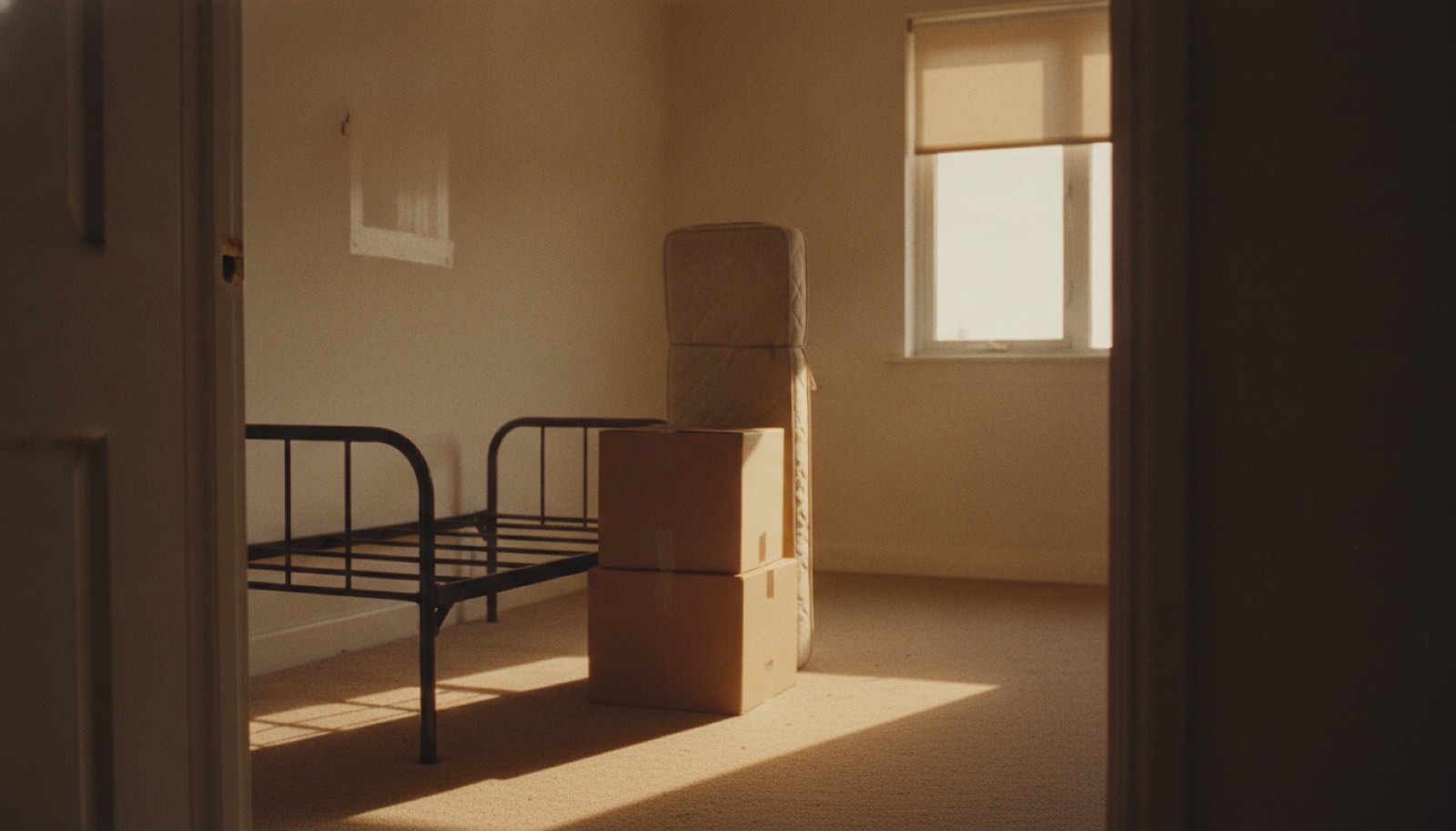 An emptied bedroom seen from the doorway, a stripped bed frame, a rolled mattress and two plain unmarked boxes standing in a band of late afternoon sun