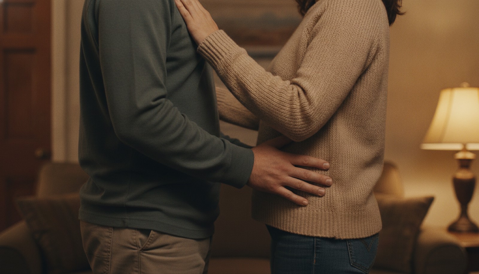 A couple cropped from the collarbone down, his hand placed stiffly on her waist and her hand on his shoulder, a small gap of air still between them in a lamp-lit living room