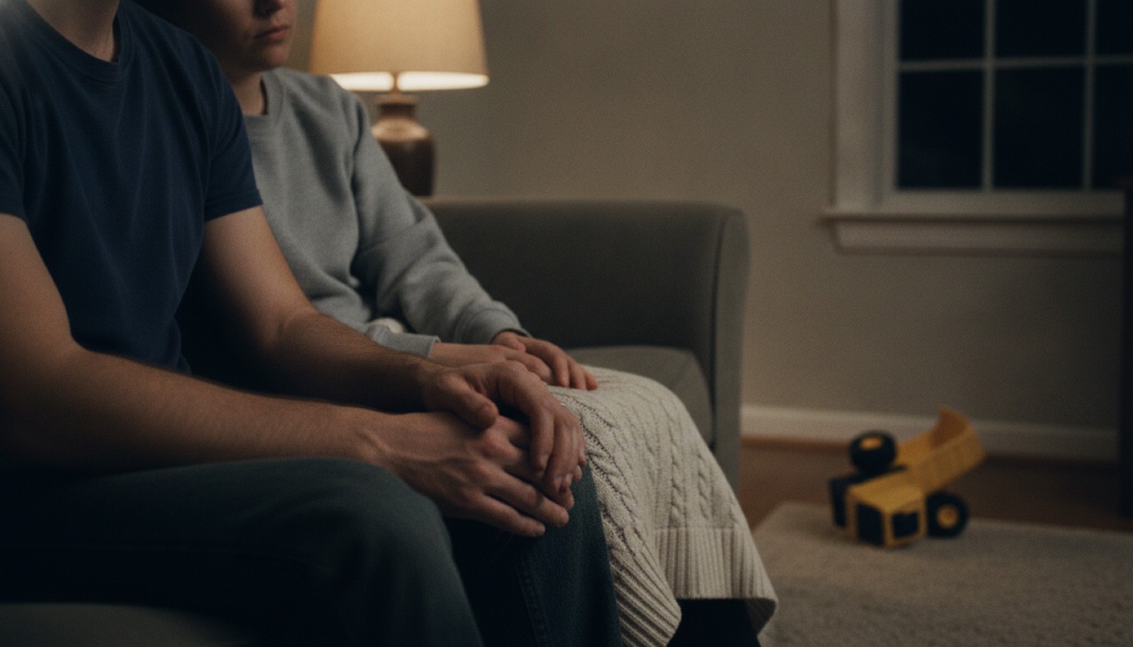 Two adults sit close together on a dim living room couch late at night with their hands joined on his knee