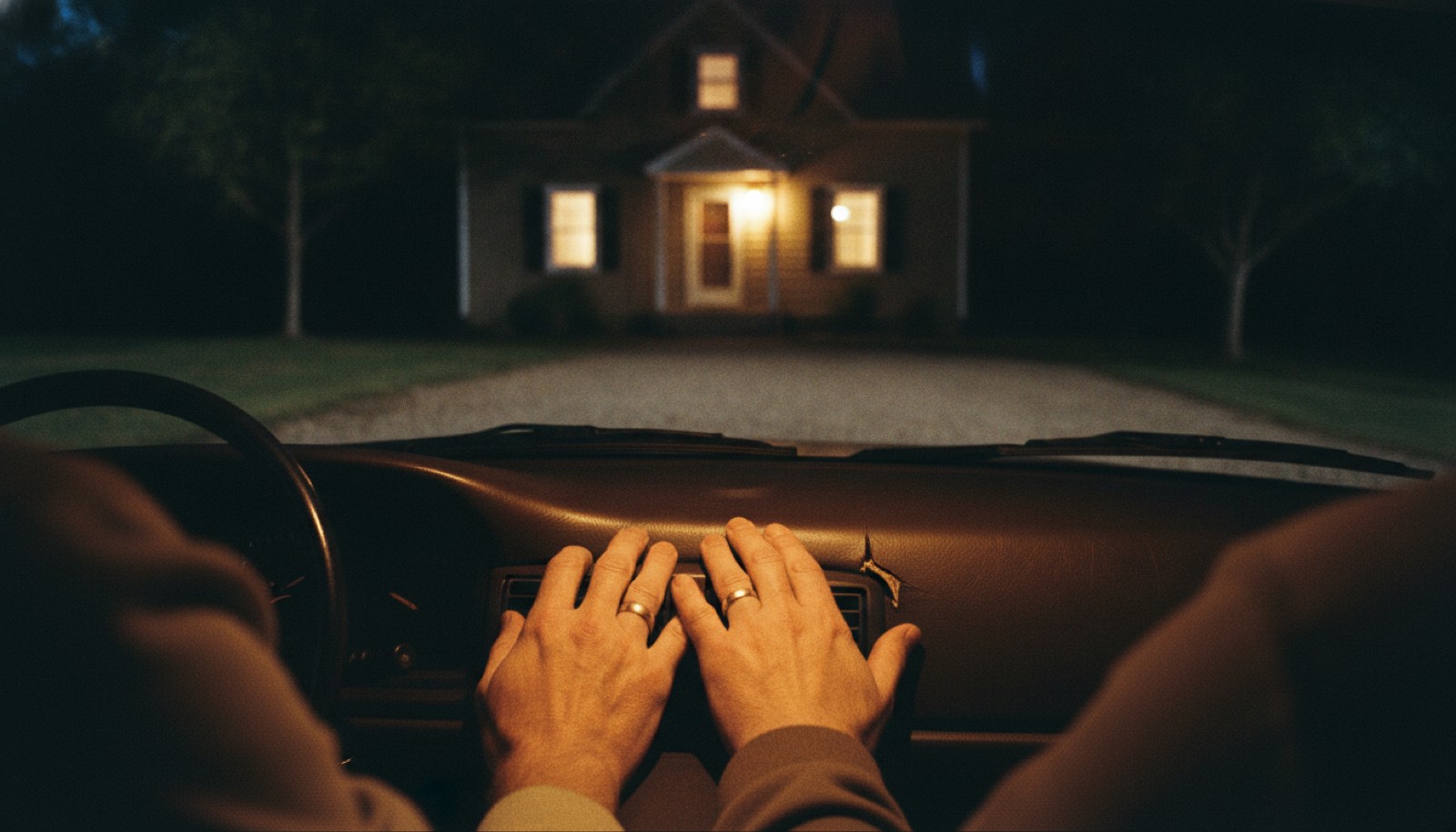 Two hands resting on a car console in a dark driveway with the house windows lit up ahead