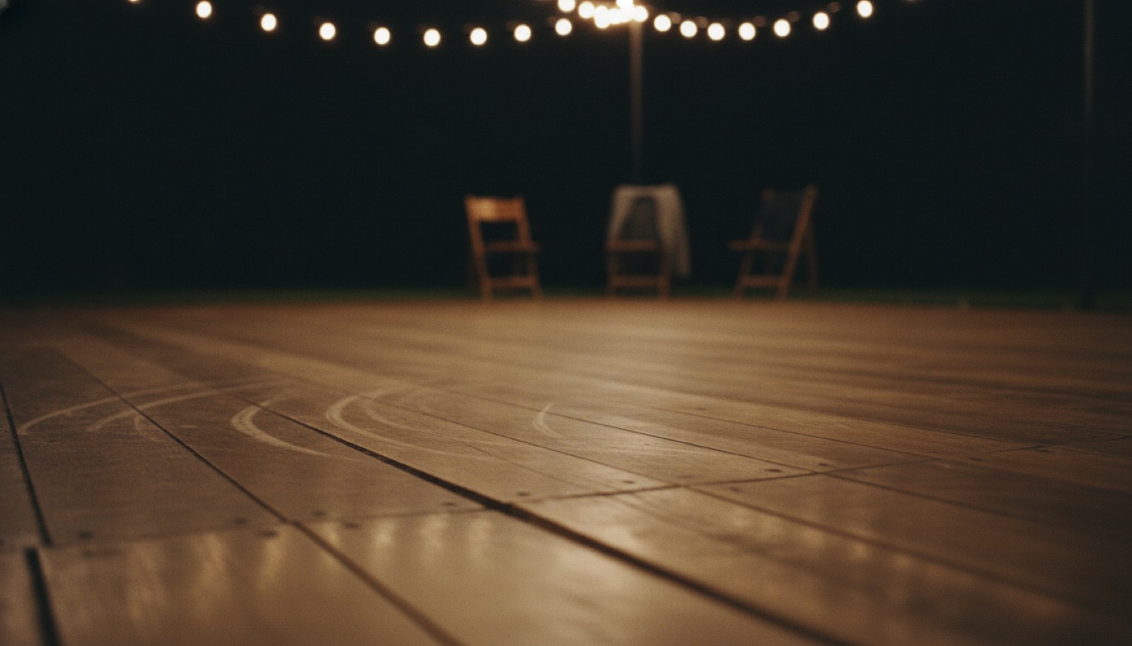 A polished wooden dance floor under warm string lights with a few empty chairs at the edge