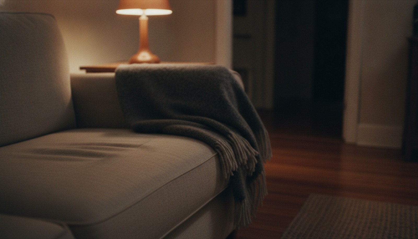 Two side by side impressions in a linen couch cushion with a folded grey wool throw over the arm, lit by one amber lamp in an otherwise empty living room