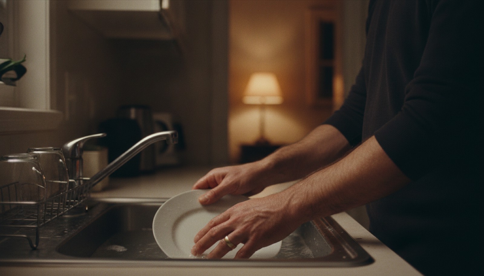 A man's hands in the dish water at a kitchen sink at night, wedding band on, with a lamp burning in the next room