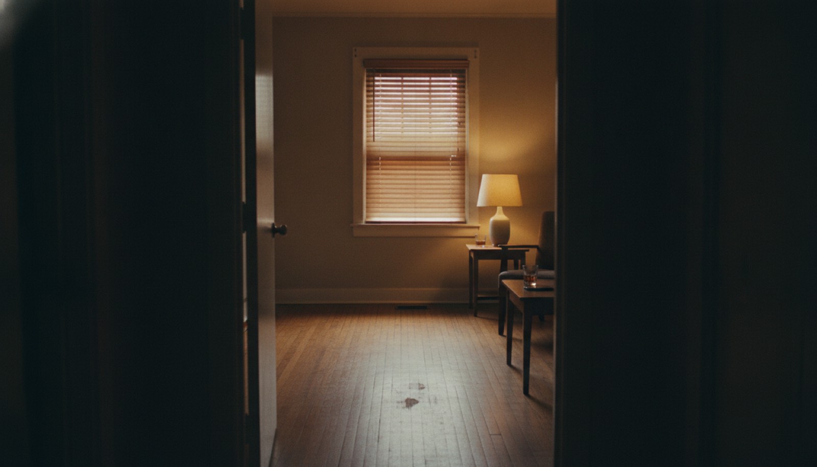 An empty room seen through a doorway from a dark hallway, the blinds pulled fully shut, one corner lamp burning low and two half-finished glasses on a side table