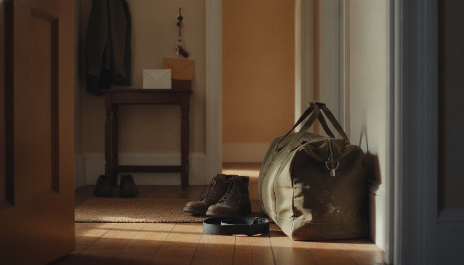 A zipped canvas duffel bag resting on a front hall floor beside two pairs of boots, with a stack of unopened mail on the hall table and late gold daylight across the boards