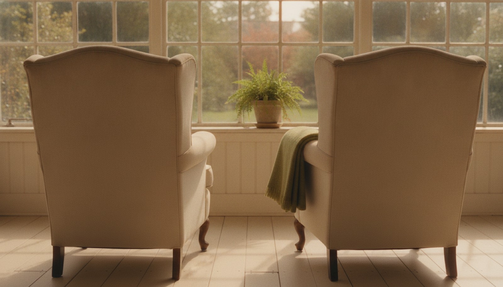Two worn armchairs facing a living room window in late morning light, one with a folded throw over the arm