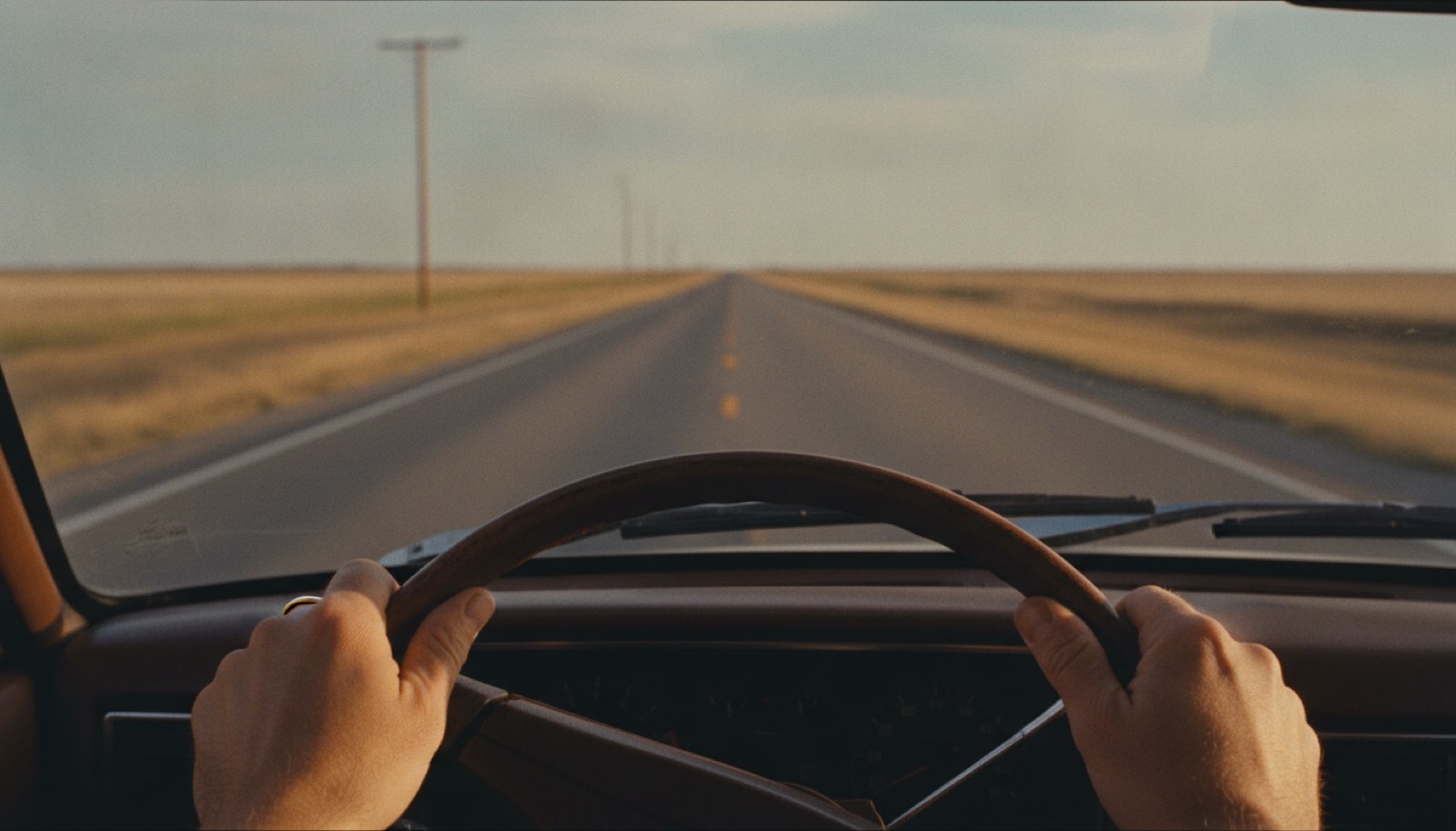 A driver's hands resting low on a worn steering wheel with an empty two-lane highway ahead in late golden light