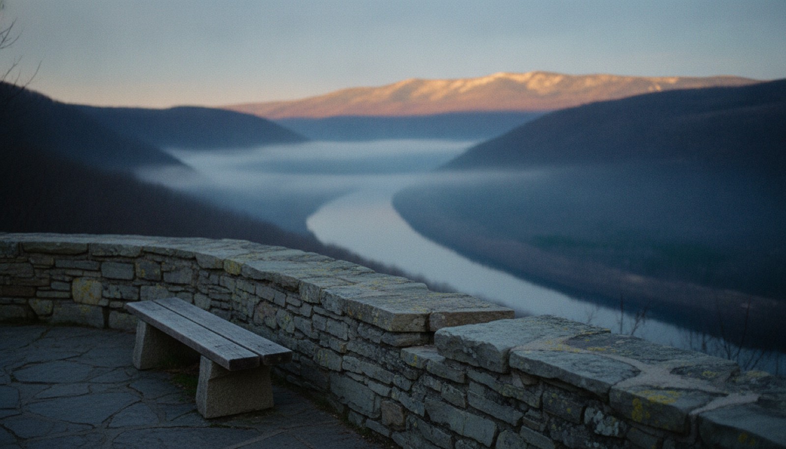 A stone overlook wall above a river valley at dusk with a single bench facing the view