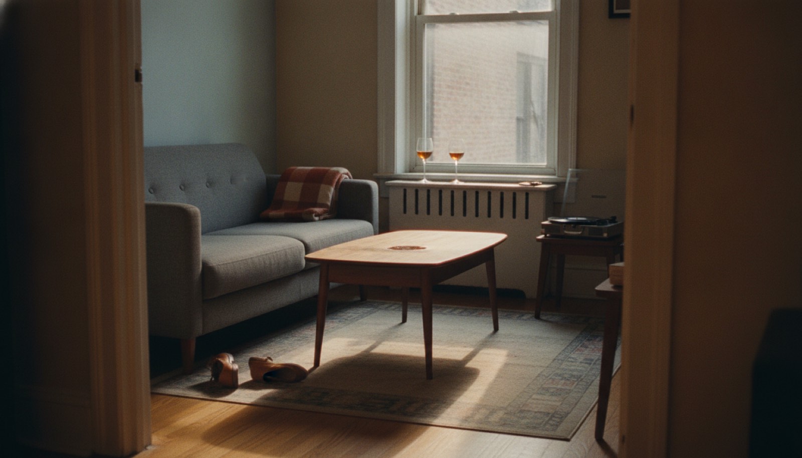 A small apartment living room in late afternoon sun with the coffee table pushed back against the couch and a record player open beside the window