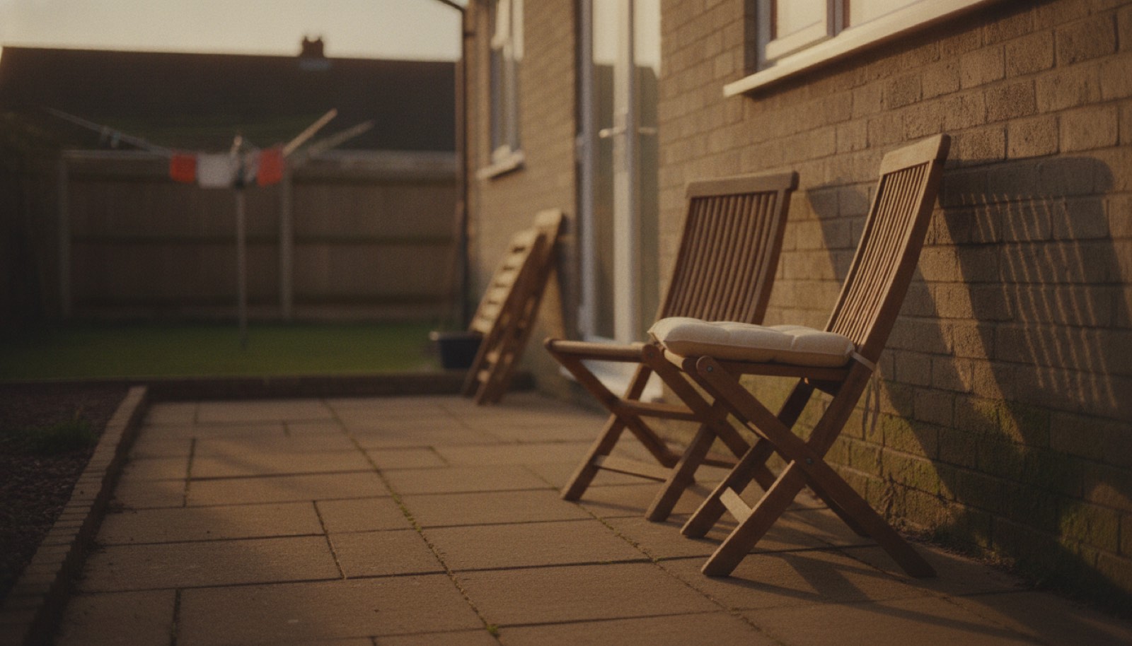 Two wooden folding chairs on a small back patio at golden hour, one opened out with a cushion on it and the other still folded flat against the house wall