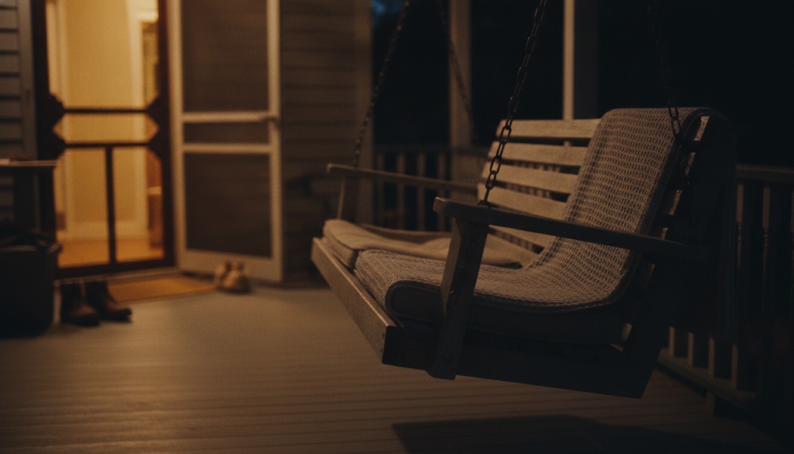 One end of a porch swing at evening with a rumpled blanket on the near seat and the far half empty, lit by warm lamplight from the open house door