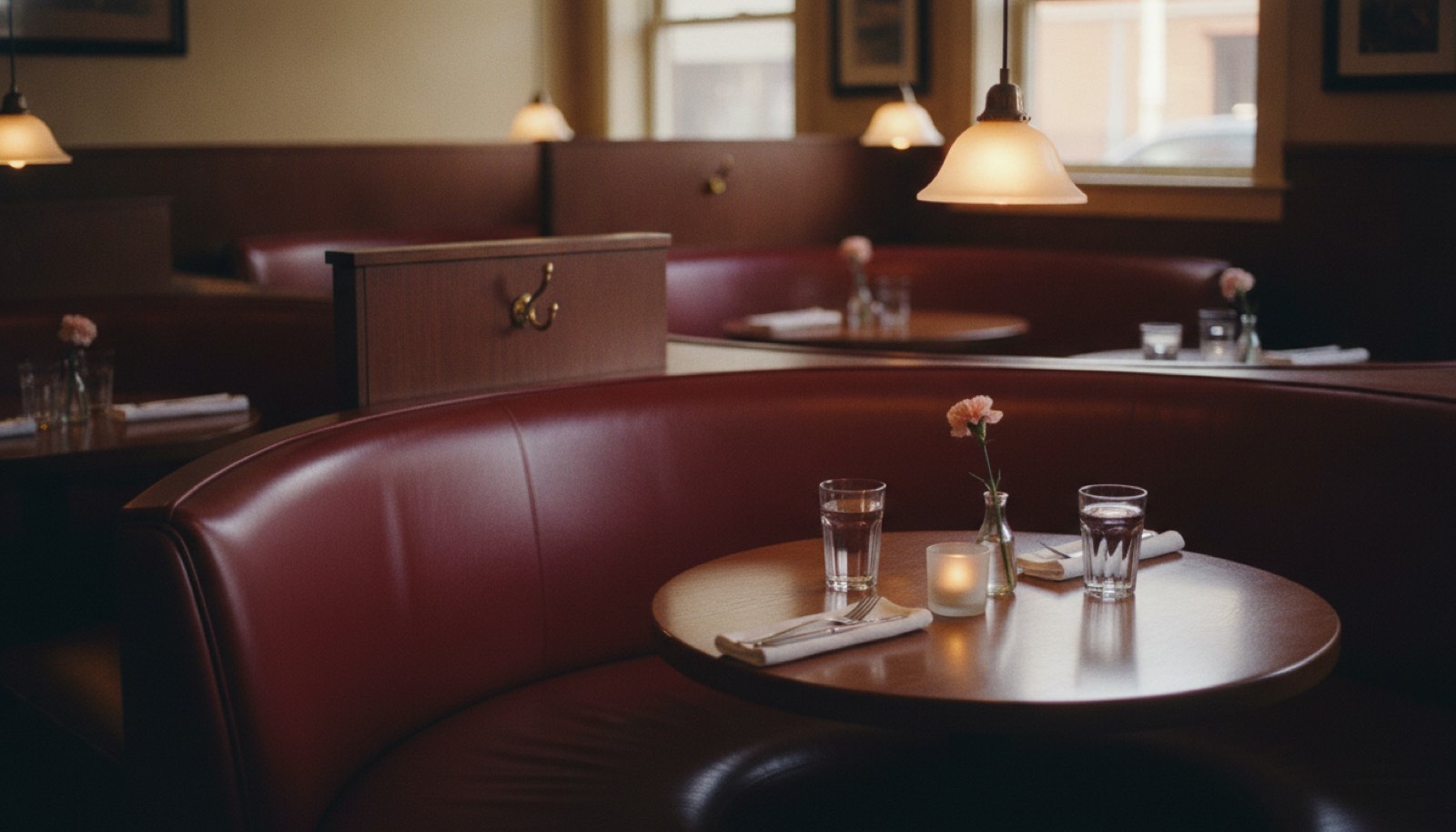 An empty curved red booth in a restaurant before the dinner crowd arrives, set for two with water glasses, a small candle and a single carnation
