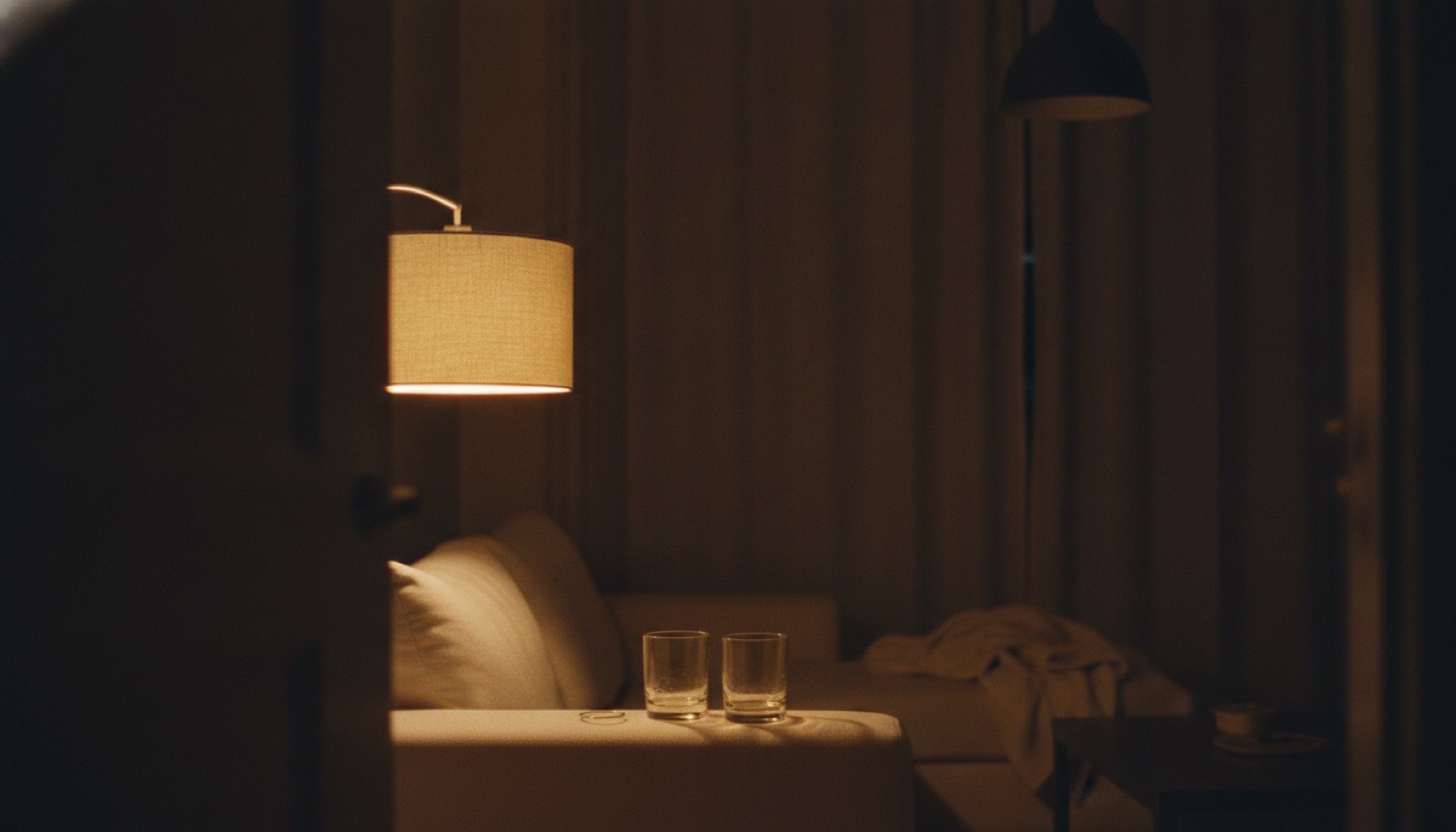 A living room lamp switched on beside two glasses on a low table, with the overhead light off and a folded blanket pushed to one end of the couch