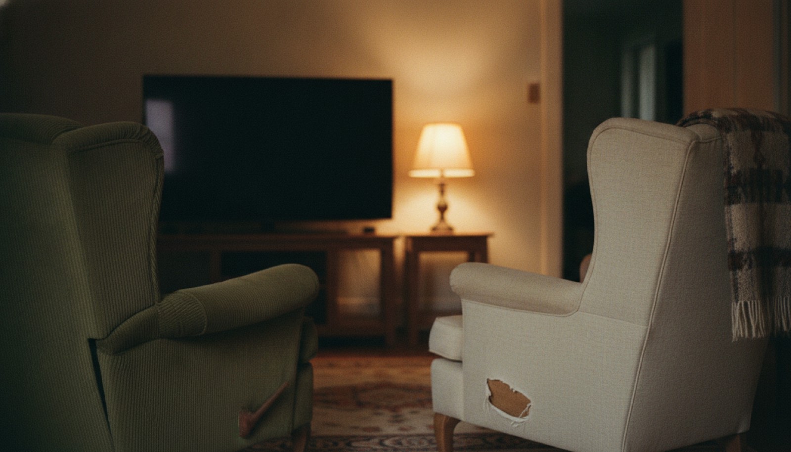 Two mismatched armchairs pulled around to face each other in a lived-in living room, with one lamp on and the television dark