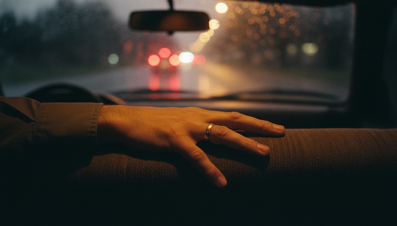 A driver's hand resting along the top edge of the empty passenger seat while stopped in traffic, with a rain flecked windscreen and blurred amber and red tail lights ahead