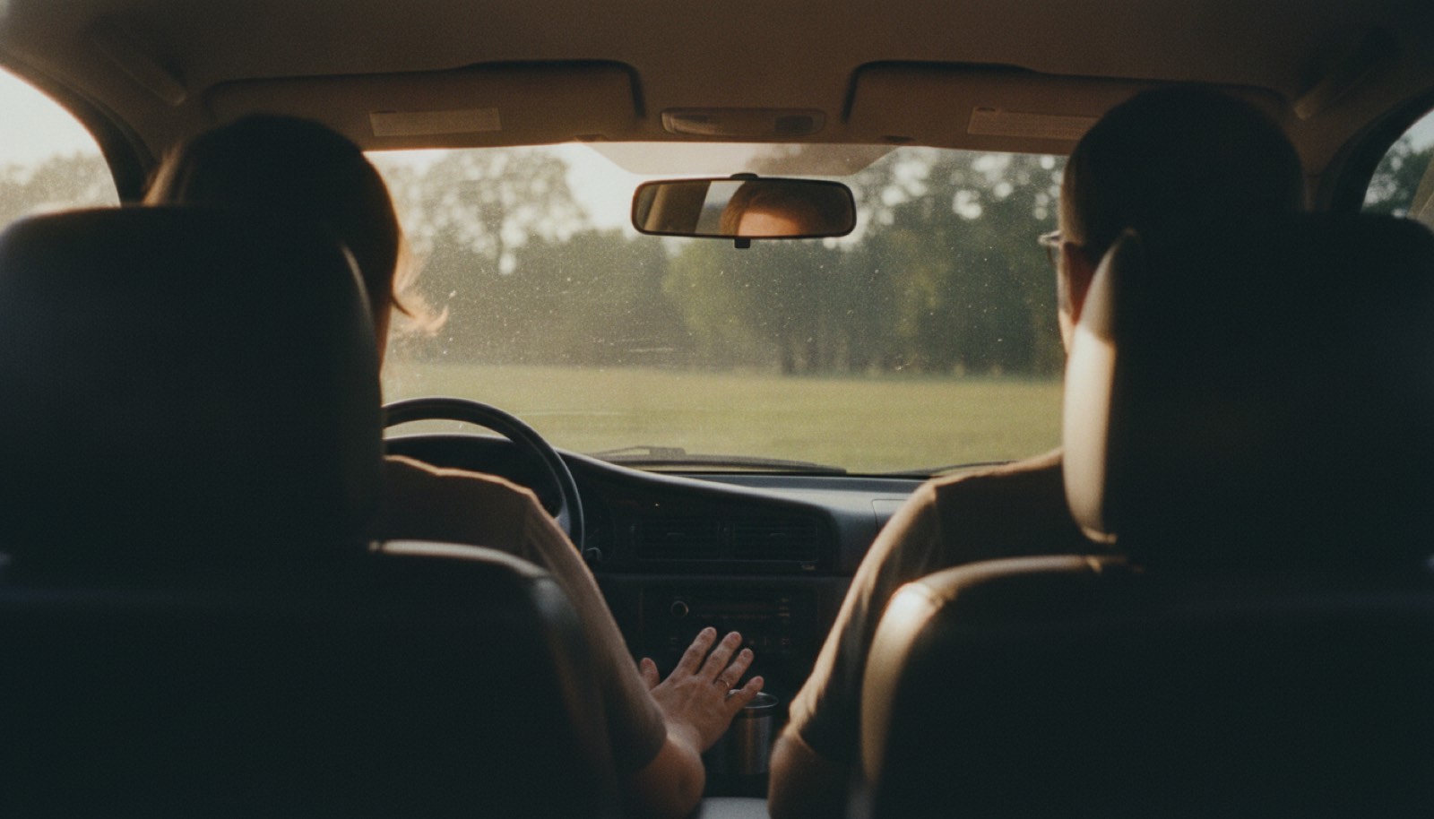 Two adults sitting in the front seats of a parked car in late afternoon sun, seen from the back seat, one hand resting on the center console