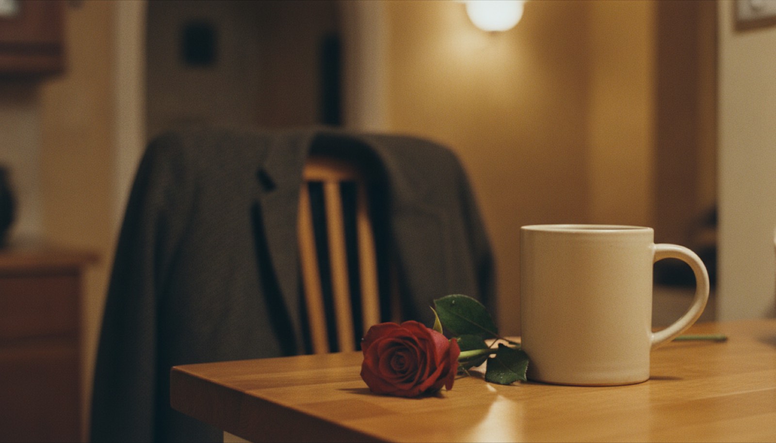 A single red rose resting on a kitchen counter beside a stoneware mug, with a wool coat over a chair in the background