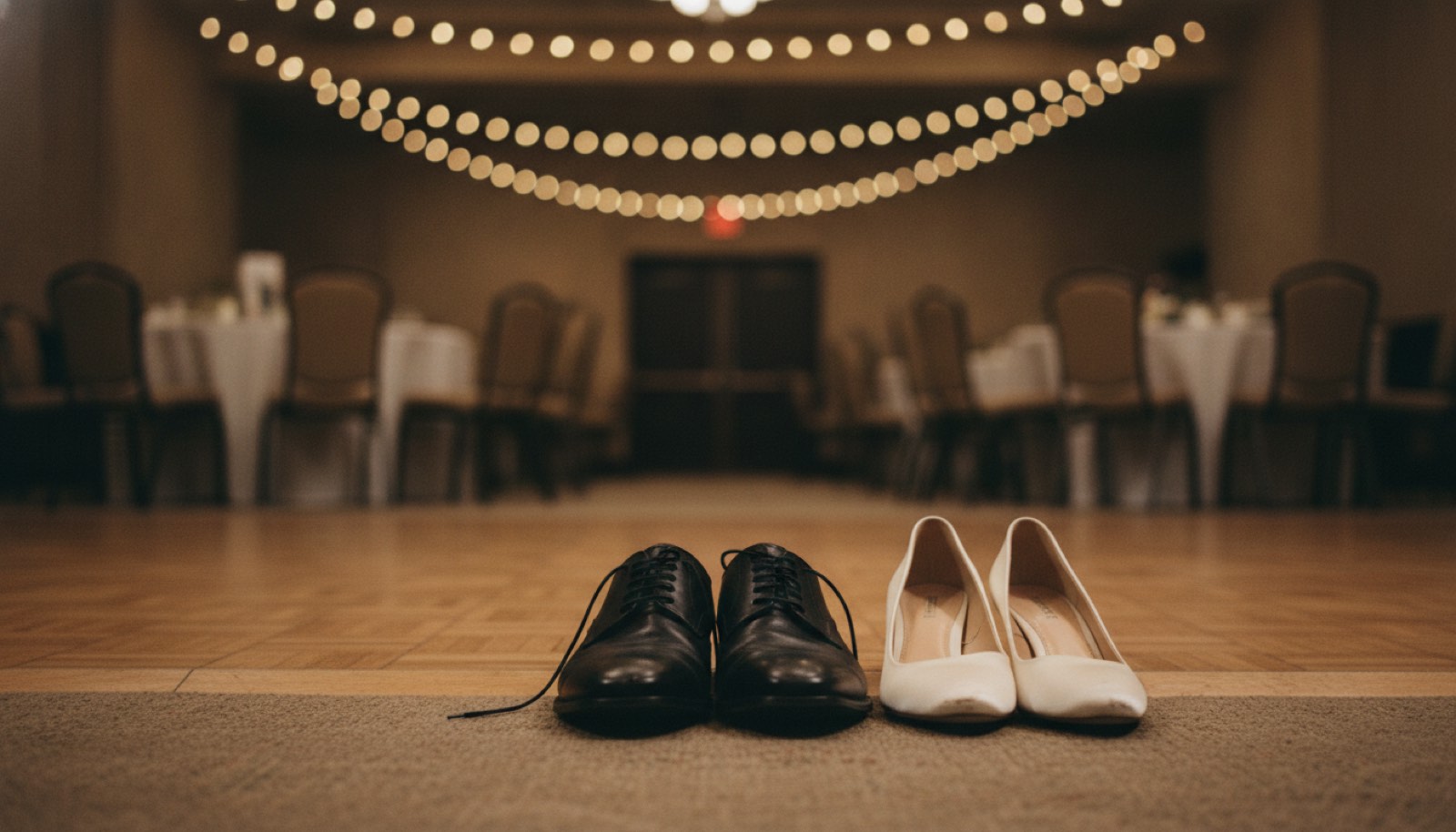 A man's leather dress shoes and a pair of low satin heels set side by side on a parquet floor at the edge of a reception dance area under strung bulbs