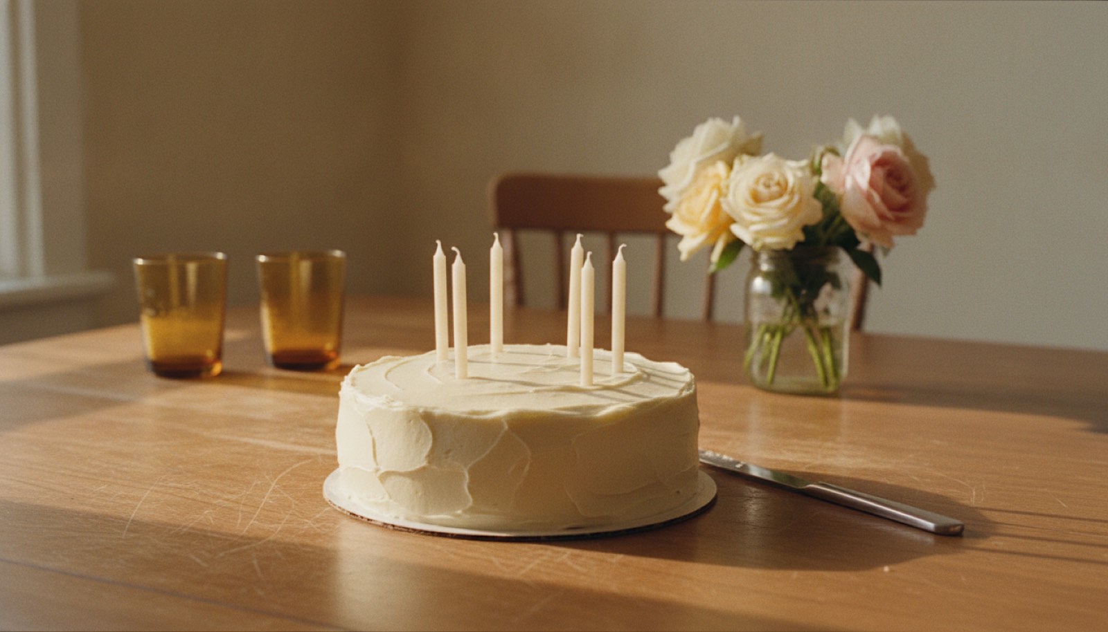 A plain ivory frosted birthday cake with six unlit candles on a scratched oak dining table beside two amber tumblers and a jar of garden roses